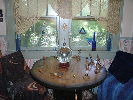 Clairvoyant psychic reader, divination specialist Lady Passion's 'div nook' for in-person psychic clairvoyant readings and divinations shows crystal ball and Tarot High Priestess symbol on a pentagram-inscribed Ouija table with two seats and a palmistry hand on the wall